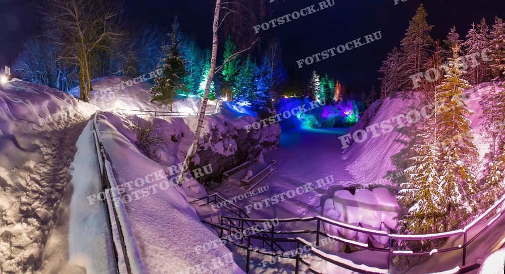 На фотографии изображён горный парк Рускеала зимой, освещённый яркими разноцветными огнями. Заснеженные тропы, скалы и деревья выглядят особенно впечатляюще под светом прожекторов, создавая сказочную атмосферу. Ночная подсветка подчёркивает контрасты и текстуры природного ландшафта, превращая его в волшебное зрелище. Это популярное туристическое место в Карелии особенно красиво в зимнее время, когда природа и искусственное освещение создают неповторимую картину.